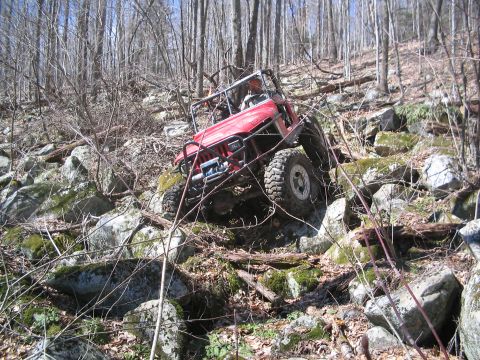 1995 Ford Bronco - A red off-road vehicle tackles a challenging rocky terrain in a forest setting.