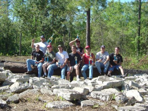 1993 Ford Bronco - A group of men pose with trophies after an event, seated on logs and surrounded by rocks in a wooded area.