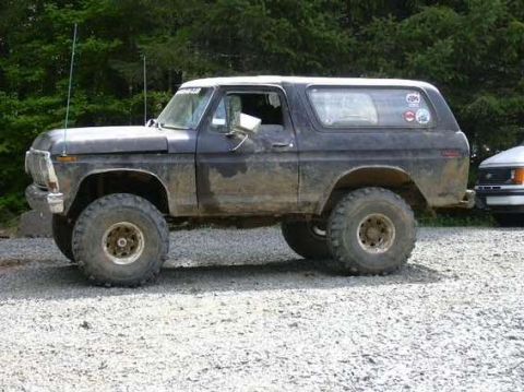 1978 Ford Bronco - A side view of a dirty 1978 Ford Bronco parked on a gravel surface with trees in the background.