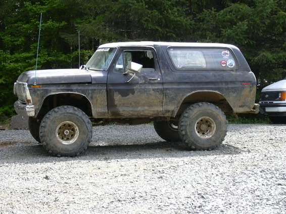 Muddy dark Ford Bronco with white top, lifted on big tires, parked on gravel.