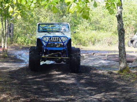 1989 Ford Bronco - A heavily modified Jeep with oversized tires navigates a muddy trail surrounded by trees.