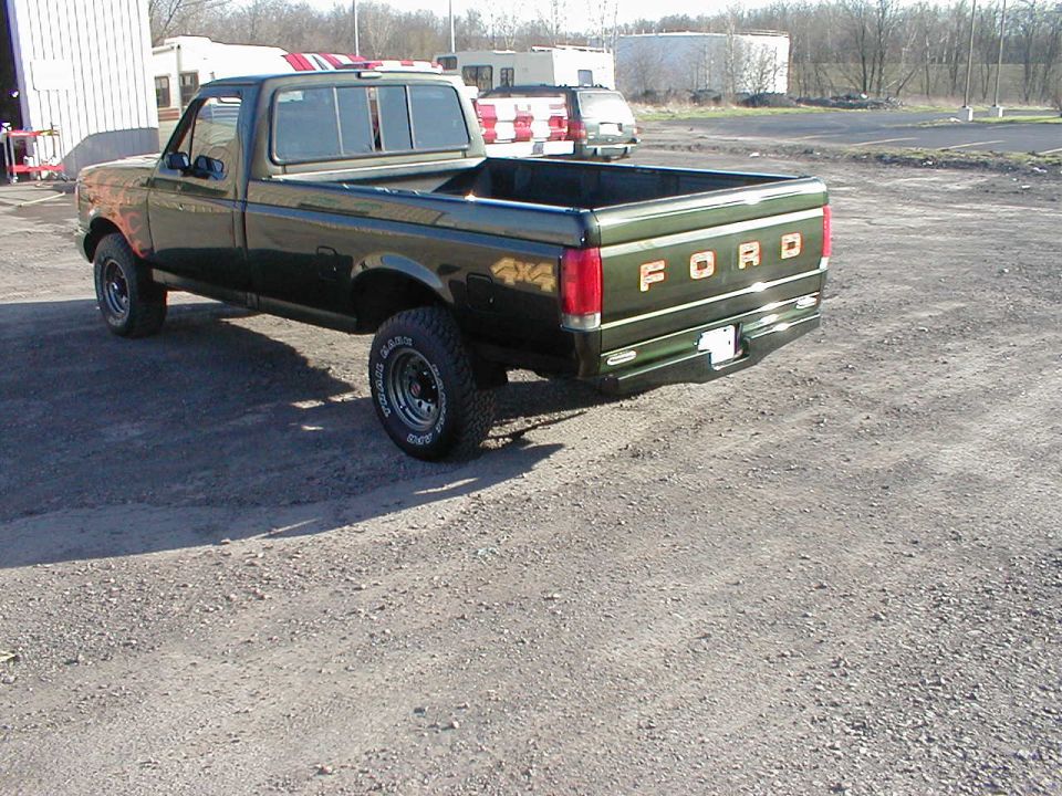 Green Ford pickup rear quarter with FORD tailgate lettering, parked on gravel lot by buildings