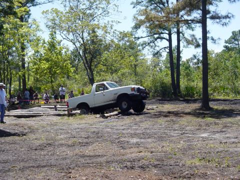 1989 Ford Bronco - A white pickup truck is shown on a challenging off-road course with spectators watching.