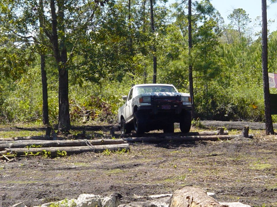 White pickup truck driving over logs in a wooded area. It has a red grille