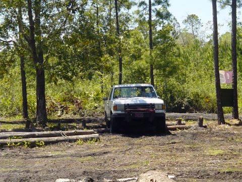 1989 Ford Bronco - A white pickup truck navigates a rough terrain course in a wooded area.