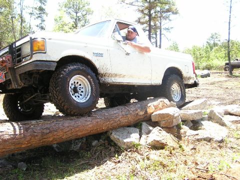 1989 Ford Bronco - A driver navigates a challenging off-road course with a white pickup truck, crossing over a large log.