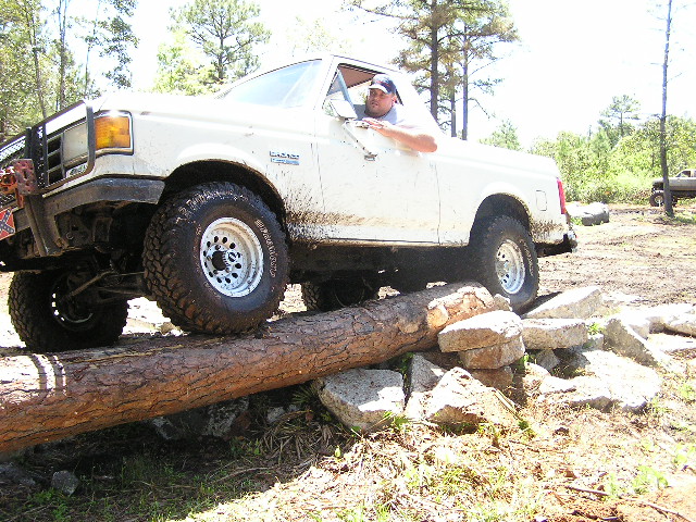 White pickup truck driving over a log obstacle. Another truck is visible in the background