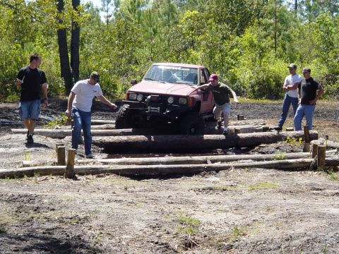 1989 Ford Bronco - A red Toyota pickup truck navigates a log obstacle course with onlookers present.