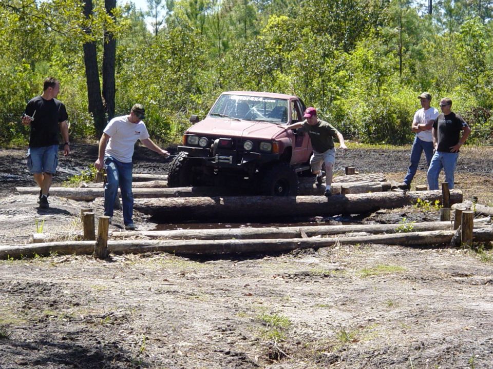 Red Toyota pickup truck with off-road modifications driving over logs, with several men watching