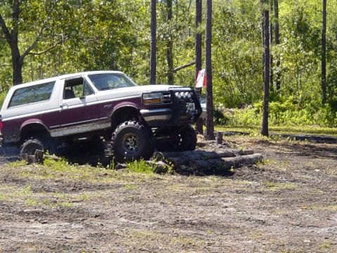 1989 Ford Bronco - Side profile of a lifted two-tone purple-over-white full-size Ford Bronco crawling over a row of logs at a wooded off-road park. It runs mu…