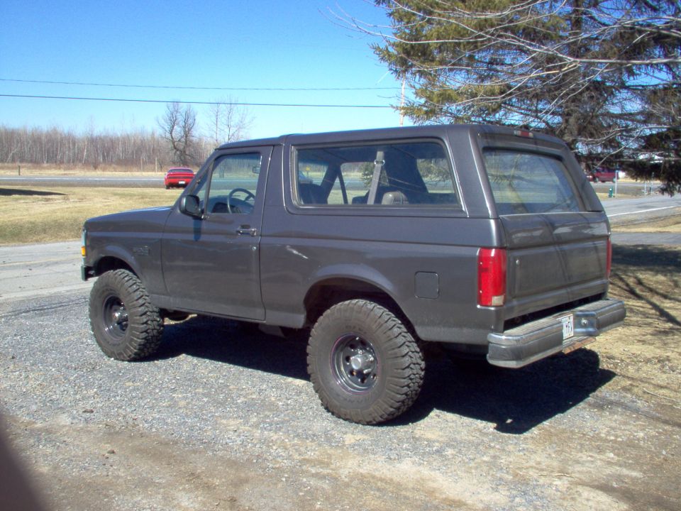 Dark gray Ford Bronco parked on gravel, rear 3/4 view. Lifted suspension, off-road tires