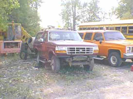 Dark red Ford Bronco without front bumper parked in grass next to an orange Suburban and school buses