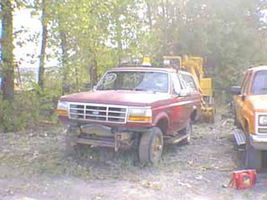 Red Ford Bronco with missing front bumper parked in a wooded gravel yard next to other vehicles
