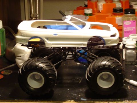 1979 Ford Bronco - A white model truck body is mounted on a monster truck chassis with oversized tires, sitting on a cluttered workbench.