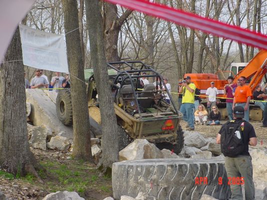 Off-road buggy with roll cage navigating a rock obstacle, spectators watch