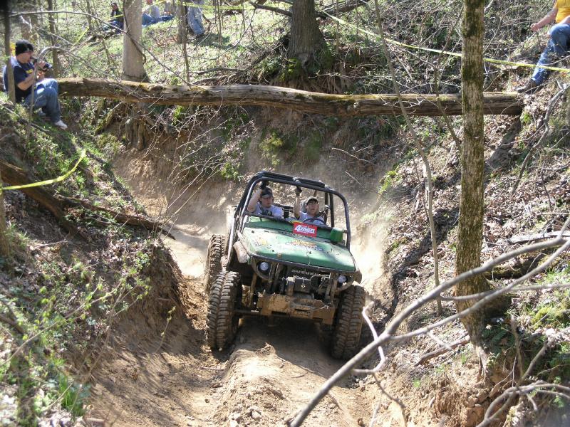 Green off-road vehicle with two people inside going up a steep dirt trail, trees and fallen log overhead