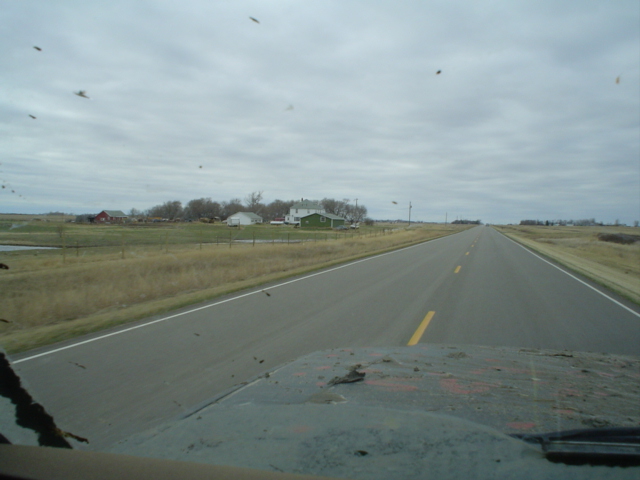 View through a windshield down a two-lane prairie highway, farmhouse and barns under gray sky