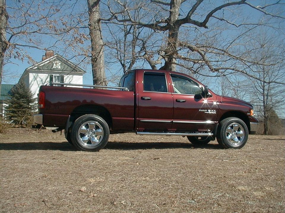 Side view of dark maroon Dodge Ram quad cab pickup with chrome wheels and side steps, parked on grass near a white house