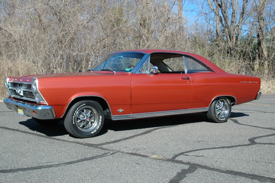 Red 1960s Ford Fairlane coupe, 3/4 front view, parked on asphalt with trees behind