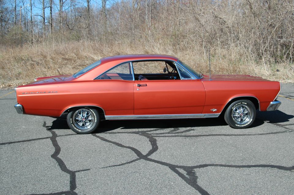 Orange 1960s Ford Fairlane hardtop coupe, side view, on asphalt with trees behind.
