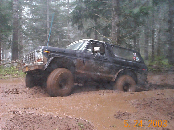 Mud-covered black and white Ford Bronco driving through a deep mud pit in the woods