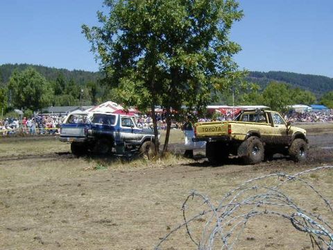 1978 Ford Bronco - Two off-road vehicles, a blue and white SUV and a yellow Toyota pickup, are seen in a muddy field with spectators in the background.