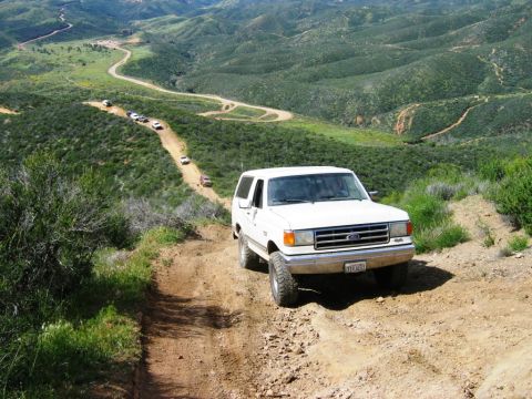 1988 Ford Bronco - A white 1988 Ford Bronco navigates a winding dirt road in a hilly, green landscape, with several other vehicles following.