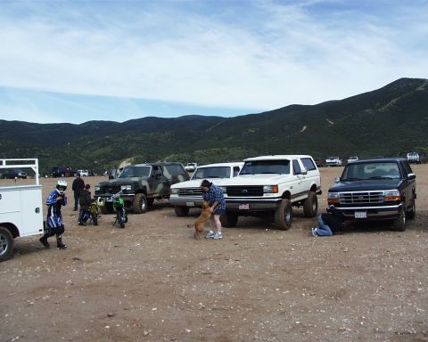1988 Ford Bronco - A group of off-road vehicles are gathered in a dusty lot with a mountainous landscape under a cloudy sky.