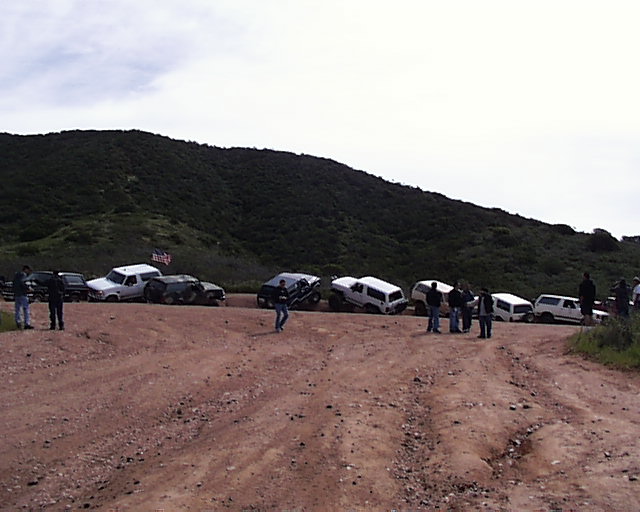 Several SUVs and trucks parked on a dirt road, some at angles, with a hill and cloudy sky