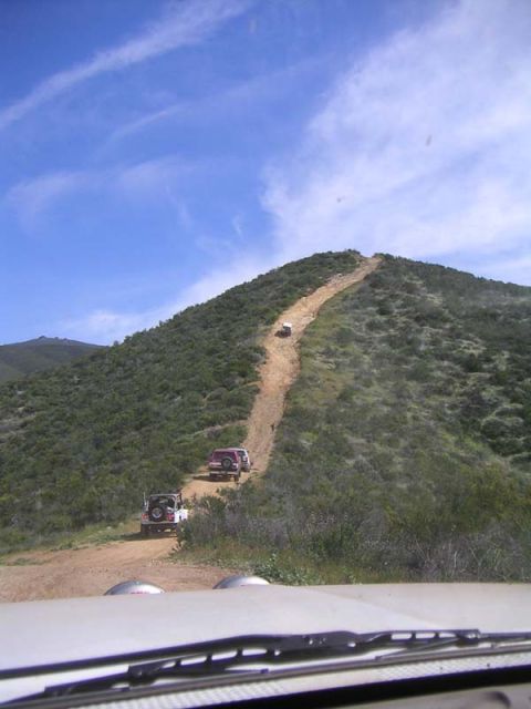 1988 Ford Bronco - Multiple off-road vehicles navigate a challenging dirt track winding up a sun-drenched hillside.