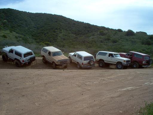 Five Ford Broncos parked on a dirt road with a grassy hill in the background