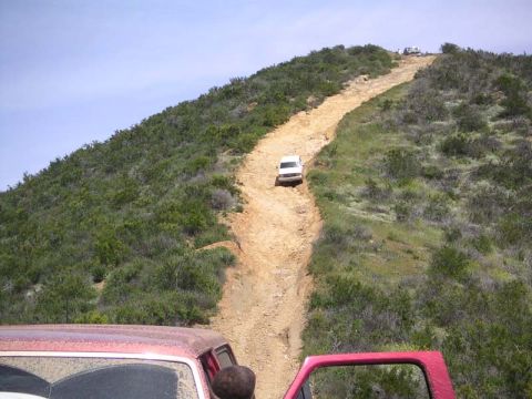 1988 Ford Bronco - Several vehicles navigate a challenging dirt path winding up a grassy, scrub-covered hillside under a clear blue sky.