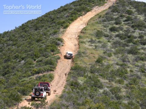 1988 Ford Bronco - Several vehicles navigate a challenging off-road trail winding up a scrub-covered hill under a clear blue sky.