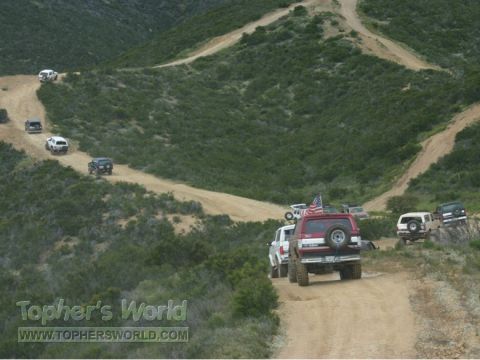 1988 Ford Bronco - Several SUVs and trucks navigate a winding dirt road through a green, hilly terrain.