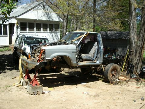 1985 Ford Bronco - A disassembled pickup truck is elevated on jack stands, with another SUV visible in the background.