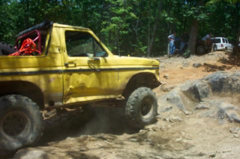 1985 Ford Bronco - A yellow SUV with significant body damage drives over rocks, with other vehicles and people in the background.