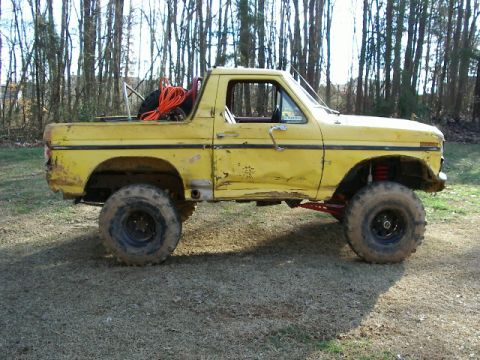 1985 Ford Bronco - This modified pickup truck sits outdoors with a backdrop of trees and a dirt ground.