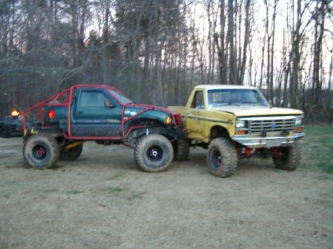 1985 Ford Bronco - Two heavily modified trucks, one yellow Ford and one green, are parked side-by-side outdoors.
