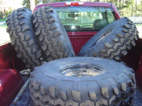 1992 Ford Bronco - Four large off-road tires are loaded into the bed of a red pickup truck.