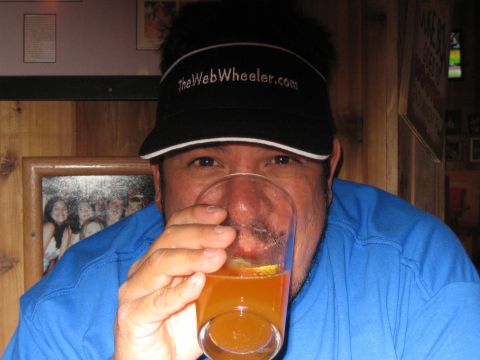 1990 Ford Bronco - man wearing a black TheWebWheeler.com visor and blue shirt drinks an orange beverage from a glass indoors.