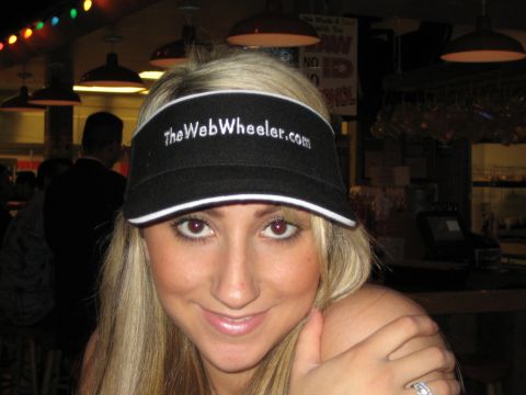1990 Ford Bronco - smiling blond woman wearing a black TheWebWheeler.com visor poses with hand on shoulder inside a lit venue.