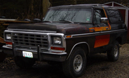 1990 Ford Bronco - brown 1970s Ford Bronco SUV with tribal side striping and chrome grille parked outdoors near a red barn.
