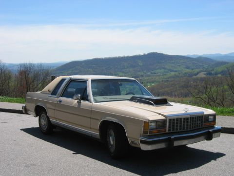 1985 Ford LTD Crown Victoria - Three-quarter front view of a tan Ford LTD Crown Victoria two-door coupe parked at a mountain overlook with green ridgelines behind. It wea…