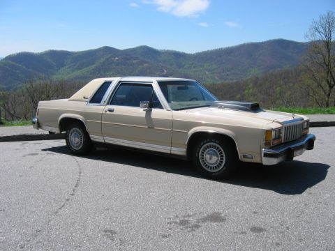 1985 Ford LTD Crown Victoria - Tan/beige two-door Ford LTD Crown Victoria parked at a mountain overlook pullout, front three-quarter view showing the vinyl-look roof with…