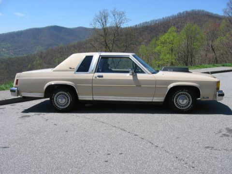 1985 Ford LTD Crown Victoria - Full side profile of a tan two-door Ford Crown Victoria coupe with a padded vinyl half-roof, opera-style rear quarter window, chrome trim,…