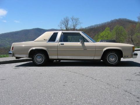 1985 Ford LTD Crown Victoria - A tan/gold two-door Ford LTD Crown Victoria coupe shown in profile (driver's side), parked on asphalt with green mountains and a blue sky b…