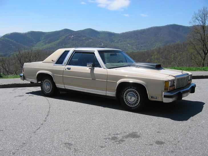 Beige two-door Ford 5.0 coupe with hood scoop parked at a mountain overlook, front three-quarter view