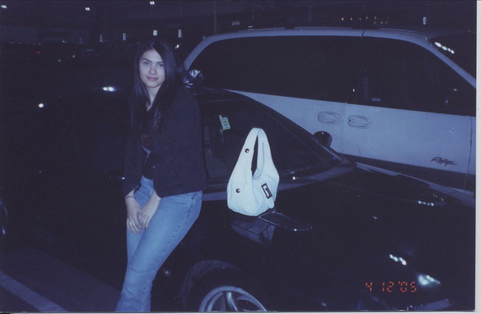 Woman in dark jacket and jeans leaning against a light-colored car in a dark parking lot