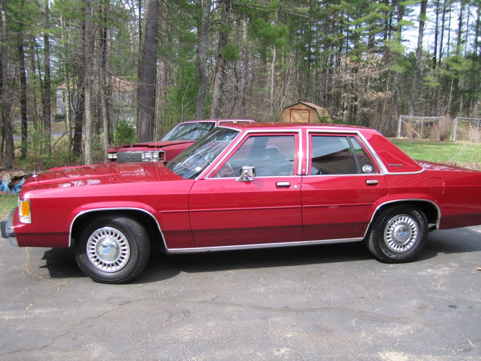 Side view of a red 1990s Ford Crown Victoria sedan parked next to another red vehicle