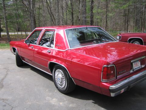 1991 Ford LTD Crown Victoria - A red sedan is parked on asphalt with trees in the background and another red vehicle partially visible.